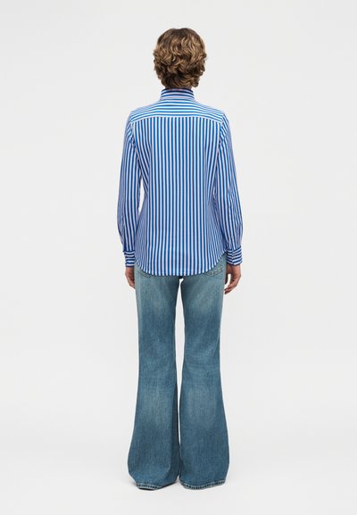 Άτομο που στέκεται με την πλάτη στραμμένη, φοράει μπλε-και-λευκό πουκάμισο με κάθετες ρίγες μακρυμάνικο και φαρδιά ξεθωριασμένα μπλε τζιν σε λευκό φόντο.