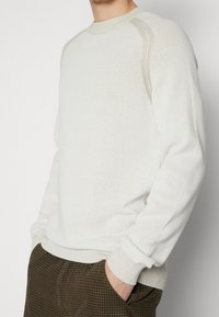 Biały dziany sweter z fakturowanym wzorem, okrągłym dekoltem i prążkowanymi mankietami. W połączeniu z brązowymi spodniami w kratę.