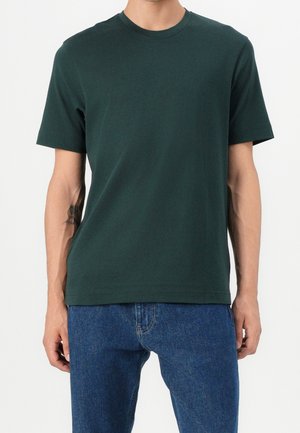 Hombre con camiseta de manga corta de cuello redondo verde oscuro y vaqueros azules, de pie frente a un fondo blanco liso.