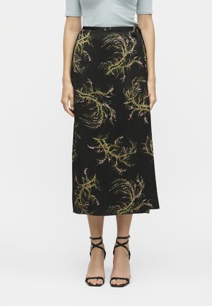 Žena oblečená v čiernej midi sukni so zelenými a ružovými kvetinovými vetvami a čiernych sandáloch na podpätku s remienkami, stojaca na bielom pozadí.