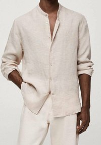 Homme portant une chemise en lin beige à boutons avec les manches retroussées et un pantalon de couleur claire, debout, une main dans la poche.