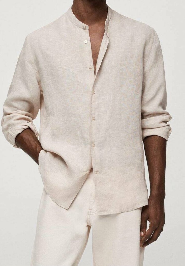 Homme portant une chemise en lin beige à boutons avec les manches retroussées et un pantalon de couleur claire, debout, une main dans la poche.