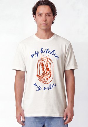 Homme portant un T-shirt blanc avec une illustration de crevette rouge et un texte bleu "my kitchen my rules" debout devant un fond blanc uni.