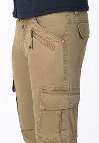 Lichtbruine cargobroek met meerdere zakken, gestructureerde stof, ritsdetails en een kleine rode logo-accent op de voorkant van de zak.