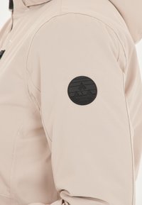 Veste beige clair en tissu lisse, dotée d'un patch circulaire noir sur l'épaule avec un logo de montagne et le nom de la marque, Whistler, Danemark.