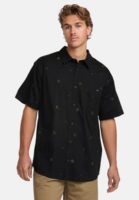 Camisa negra de manga corta hecha de tela ligera, con patrones geométricos dorados, botones en el frente y un bolsillo en el pecho.