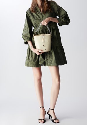Crèmekleurige emmervormige tas met krokodillenstructuur, voorzien van handgrepen van doorzichtige kunststof kettingen en een trekkoordsluiting, vormt een contrast met de kaki jurk.