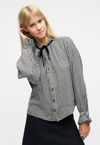 Femme blonde touchant ses cheveux, vêtue d'un chemisier à carreaux noir et blanc avec un ruban noir noué et une jupe noire, sur un fond blanc uni.