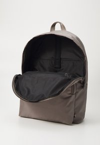 Rucsac din material maro, cu un buzunar frontal cu fermoar, interior negru spațios și o mânere de sus pentru transport ușor. Prezintă un design elegant și minimalist.