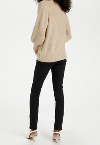 Pull en maille beige avec texture côtelée, coupe décontractée et manches longues, associé à un jean noir moulant et des talons blancs.