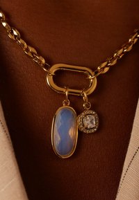 Collier en or avec un grand bijou ovale bleu à surface facettée et une petite pierre carrée transparente entourée de petits cristaux.
