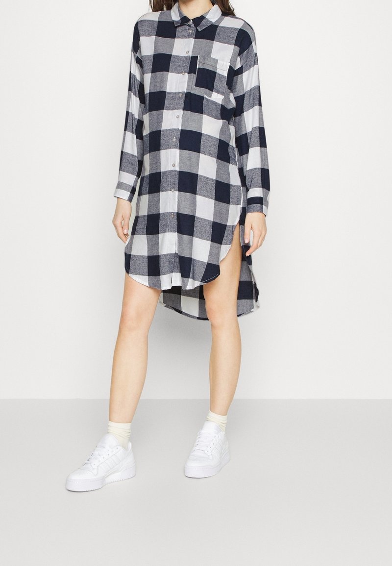 Femme portant une robe-chemise à manches longues à carreaux noirs et blancs avec une poche avant, des chaussettes blanches et des baskets blanches, debout devant un fond blanc.