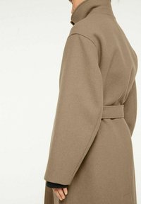 Brun, lång rock med hög krage och skärp i midjan. Har vida ärmar och en mjuk textur, visad från sidan.