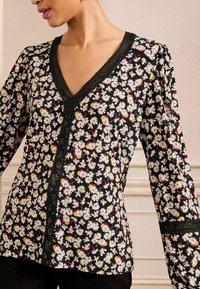 Blusa floreale con sfondo nero decorato con margherite bianche, accenti rosa e gialli, scollo a V e rifiniture testurizzate nere su maniche e parte frontale.