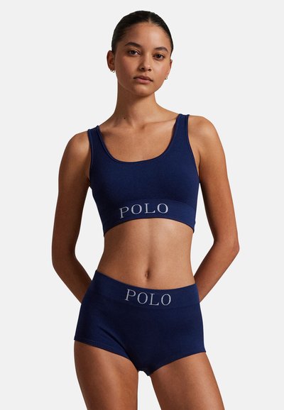 Kobieta ubrana w granatowy prążkowany krótki top i dopasowane szorty z logo "POLO" na pasku, stojąca z rękami za plecami.