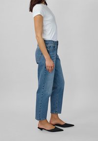 Vit ribbad t-shirt med kort ärm i kombination med högmidjade blå jeans och spetsiga svarta mules. Neutral bakgrund för att fokusera på outfiten.