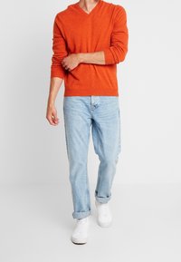 En orange v-ringad tröja i lättstickad textur, matchad med ljusblå jeans. Vita sneakers fulländar outfiten. En enkel look mot en enfärgad bakgrund.
