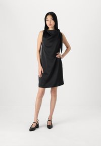 Svart ärmlös klänning med ett flödande mönster, en draperad halsringning och en sidobåge som accent. Slät tyg, knälång siluett och minimal textur.