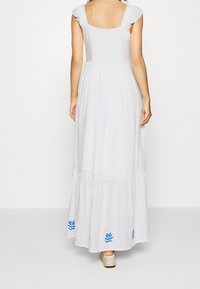 Vrouw draagt een witte mouwloze maxi-jurk met blauwe bladpatronen bij de zoom, staat blootsvoets tegen een effen witte achtergrond.