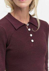 Burgundy ribbed knit top dengan kerah, menampilkan lima aksen tombol perak dan desain yang pas. Tekstur lembut dan bentuk terstruktur.