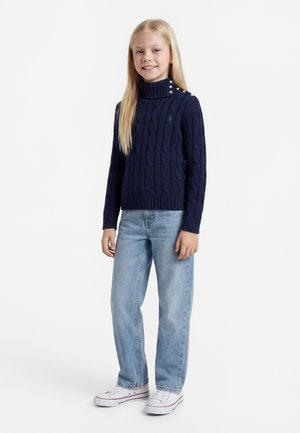 Mädchen mit langen blonden Haaren, das einen marineblauen, kabelgestrickten Rollkragenpullover, hellblaue Jeans und weiße Turnschuhe trägt, steht und lächelt.