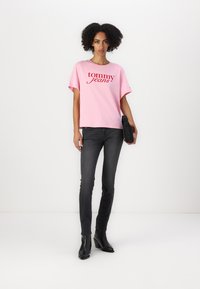 Rosa bomull t-skjorte med "tommy jeans" logo i rødt, kombinert med smale svarte jeans og svarte ankelstøvler, satt mot en hvit bakgrunn.