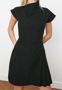 Une femme porte une robe noire élégante avec des manches courtes et une jupe évasée, dotée de poches, devant un fond uni.