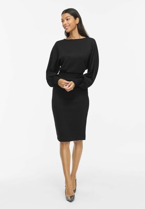 Un mannequin portant une robe noire longueur genou à longues manches bouffantes et des escarpins noirs pointus se tient debout, les mains jointes, sur un fond blanc.