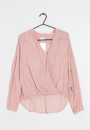 Blusa de manga larga de color rosa claro con patrón de puntos rojos, escote en V, cintura fruncida y parte trasera ligeramente más larga, colgando de una percha negra sobre fondo blanco.