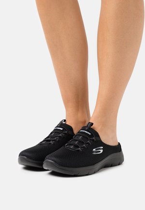 Fødder iført sorte slip-on Skechers sneakers med elastiske snøredetaljer og en hvid "S"-logo på siden mod en ensfarvet baggrund.