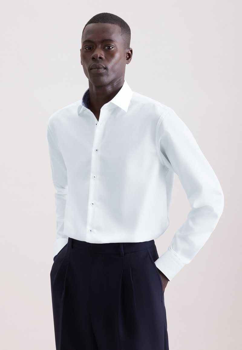 Homme à la peau foncée portant une chemise blanche à manches longues boutonnée, rentrée dans un pantalon taille haute foncé, debout les mains dans les poches.