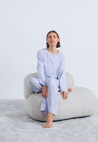 Conjunto de ropa de estar en casa de color lila claro, con mangas largas y pantalones, hecho de tela suave, usado mientras la persona está sentada en una silla beige texturizada.