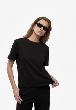 Mujer con gafas de sol negras, pendientes de aro, camisa de manga corta negra y pantalones negros, sujetando el dobladillo de su camisa con una mano.