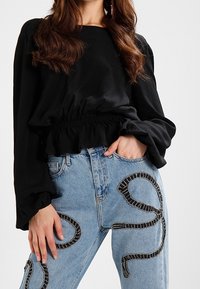 Black long-sleeve blouse with a gathered waist, paired with light blue denim jeans featuring decorative black and silver stitching patterns.