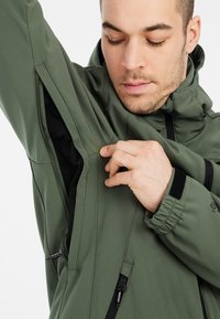 Grøn softshell jakke med høj krave, justerbare manchetter og lynlåset ventilation under armene. Har et sømløst design og struktureret stof.