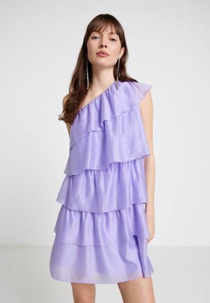 Giovane donna che indossa un abito lilla senza maniche con tre balze sovrapposte e lunghi orecchini pendenti di cristallo su uno sfondo semplice.