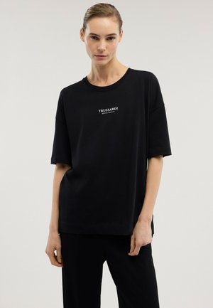 Frau trägt ein lockeres schwarzes T-Shirt mit dem weißen Schriftzug "TRUSSARDI GENTLE SOCIETY" und steht vor einem einfarbigen hellen Hintergrund.