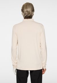 Langärmliges Poloshirt aus leicht beigem Stoff mit lockerer Passform, flachem Kragen und gerippten Bündchen. Die Rückansicht zeigt eine glatte Textur.