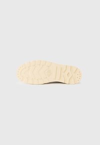 Suela de zapato con una superficie de goma beige texturizada, que presenta un patrón de relieve en zigzag y pequeños agujeros de drenaje circulares.
