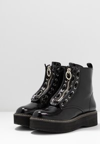 Bottes plateforme en cuir verni noir avec des zips à l'avant, des tirettes circulaires en argent et des œillets décoratifs en métal sur les côtés.