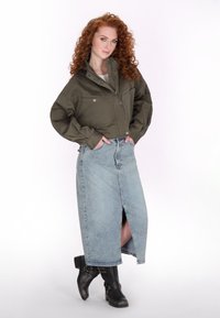 Olijfgroene cropped jack met knoopdetails, gecombineerd met een lichtblauwe denim maxi rok met een vooropening. Zwarte enkellaarzen maken de look af.