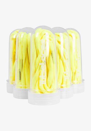 Žluté ploché tkaničky balené v průhledných plastových trubicích, s viditelným brandingem a texturovaným povrchem. Každá trubice obsahuje více délek tkaniček.