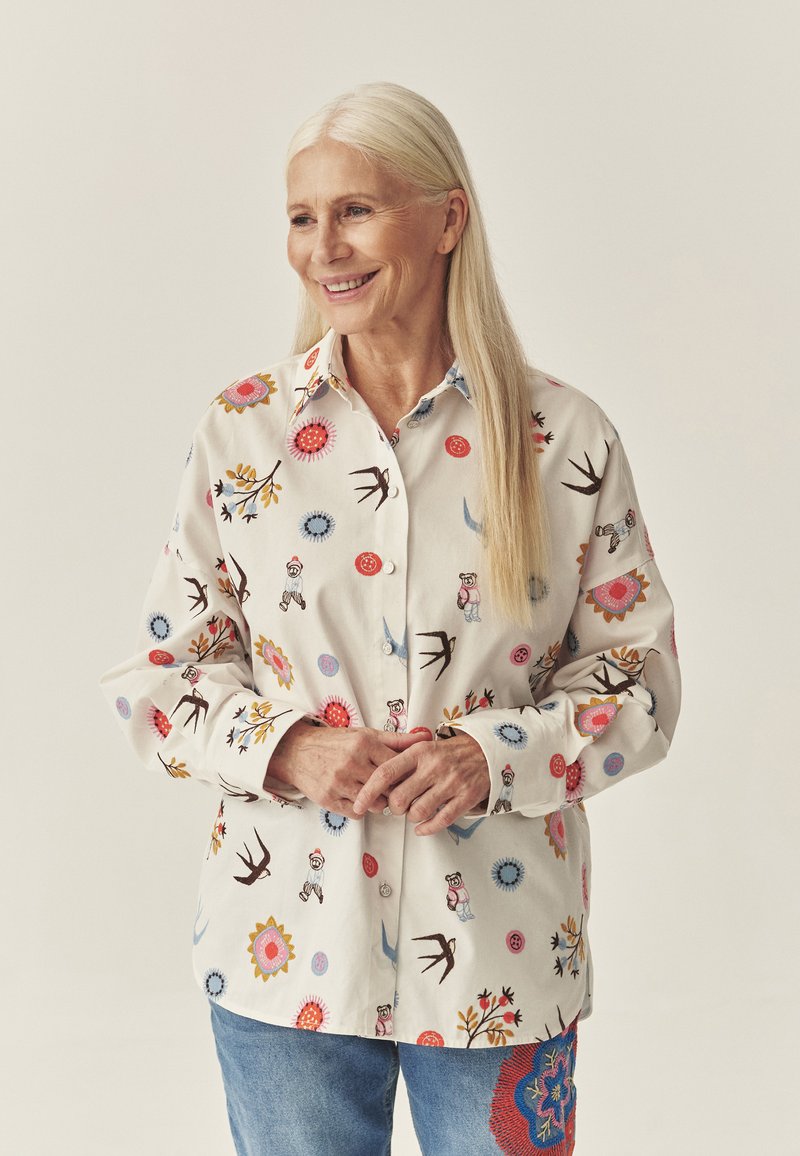 Valge puuvillane särk, millel on värvikas lille- ja loomade tikand, ees nööpidega sulgur ja kraega kaelus. Sobitatud siniste teksadega.