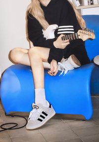 Bílo-černé tenisky Adidas se třemi černými pruhy, doplněné o bílé ponožky, na pozadí modré zakřivené židle. Kytara je držena uvolněně.