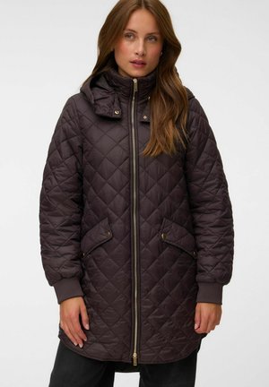 Femme portant une longue veste matelassée marron foncé avec une fermeture éclair sur le devant, des poches à boutons-pression et un col montant avec une capuche.