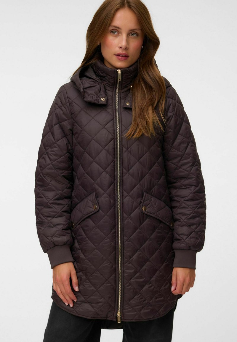 Femme portant une longue veste matelassée marron foncé avec une fermeture éclair sur le devant, des poches à boutons-pression et un col montant avec une capuche.