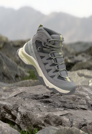 Grå og beige tursko med Gore-Tex-stoff, snøringen knyttet, hengende over steinete terreng utendørs med uklart fjellandskap i bakgrunnen.