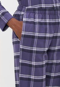 Mano descansando dentro del bolsillo de un pantalón de cuadros morados con una cinturilla elástica, también es visible la manga de una camisa de cuadros a juego.
