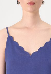 Top blu senza maniche con scollatura a onda, realizzato in tessuto leggero. Presenta spalline sottili e una texture morbida.