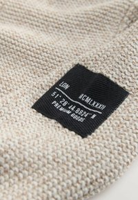 Bézs színű szőtt textil durva textúrával; tartalmaz egy fekete címkét fehér szöveggel, földrajzi koordinátákkal és márkanévvel.